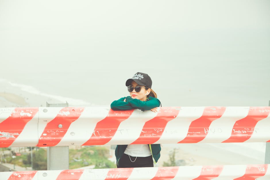 A woman in sunglasses and a cap leans on a red and white barrier by the foggy seaside.